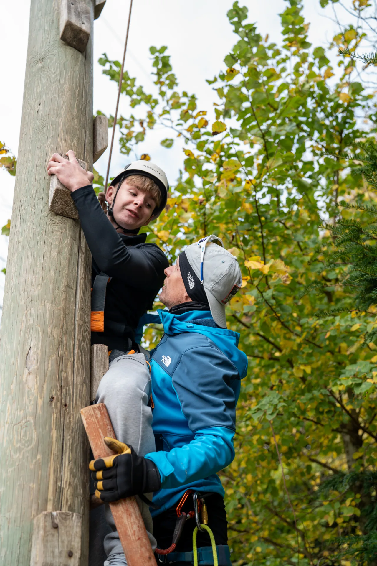 Two climbers navigate a vertical pole in a forested setting, demonstrating teamwork and physical endurance.